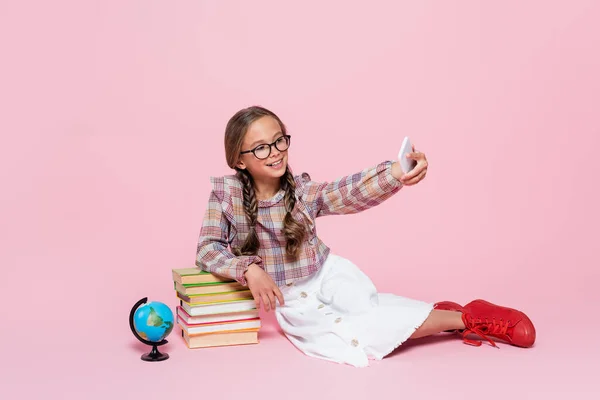 Smiling girl taking selfie while sitting near stack of books and globe on pink background — Photo de stock