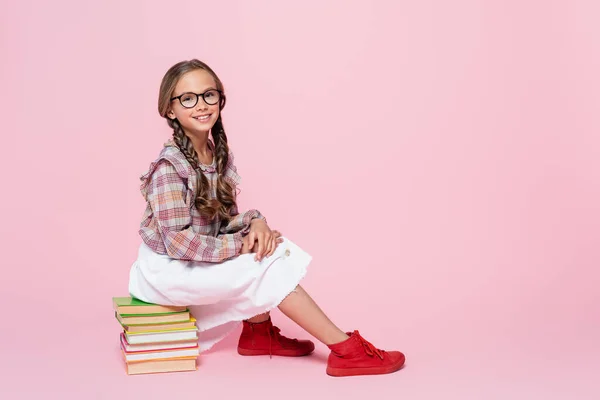 Stylish child smiling at camera while sitting on stack of books on pink background — Photo de stock