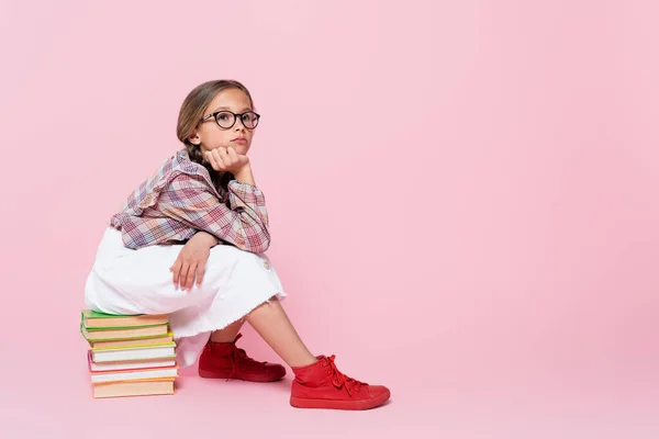 Ragazza pensierosa in camicetta a quadri e gonna bianca seduta sui libri e distogliendo lo sguardo sullo sfondo rosa — Foto stock