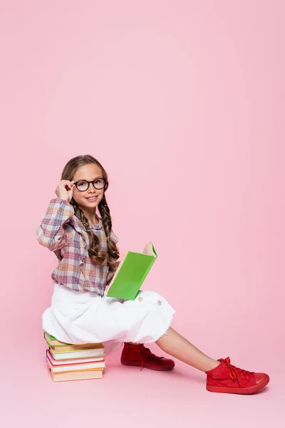 Fröhliches Kind, das Brillen justiert, während es auf Lehrbüchern auf rosa Hintergrund sitzt — Stockfoto