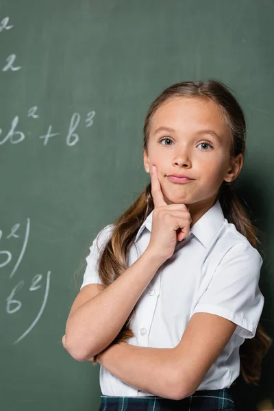 Thoughtful schoolkid looking at camera near chalkboard in school — Stock Photo