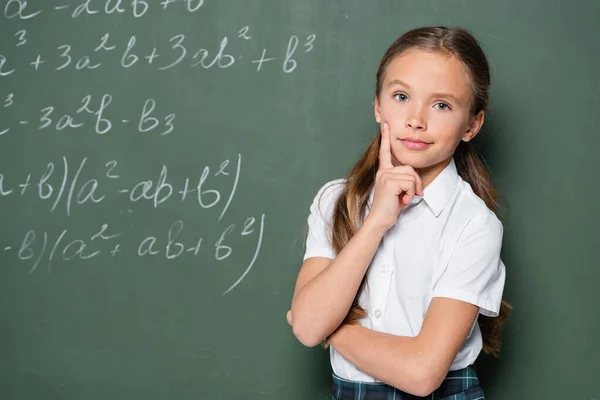 Pensive schoolgirl with hand near face looking at camera near chalkboard with equations — Photo de stock