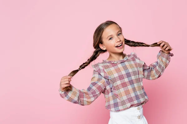 Cheerful girl in plaid blouse touching pigtails while looking at camera isolated on pink - foto de stock