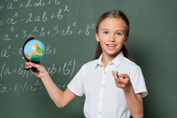 Scolaro felice con piccolo globo che punta con la matita alla macchina fotografica vicino alla lavagna con equazioni — Foto stock