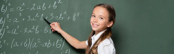 Schulmädchen lächelt in die Kamera, während sie auf Gleichungen auf der Tafel zeigt, Banner — Stockfoto