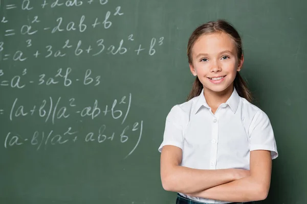 Preteen schoolkid with crossed arms smiling at camera near equations on chalkboard - foto de stock