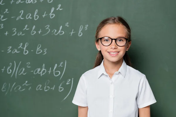 Smiling schoolgirl in eyeglasses looking at camera near chalkboard with equations — Stock Photo