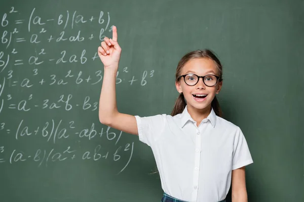 Aufgeregtes Schulmädchen mit Brille zeigt Ideengeste neben Kreidetafel mit Gleichungen — Stockfoto
