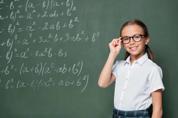 Fröhliches Schulkind justiert Brille neben Tafel mit mathematischen Gleichungen — Stockfoto