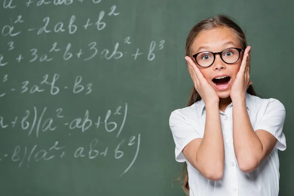 Astonished schoolchild touching face and looking at camera near chalkboard — Photo de stock