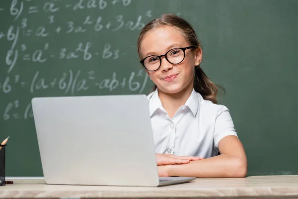 Scolaretta in occhiali sorridente alla fotocamera vicino laptop e lavagna su sfondo sfocato — Foto stock