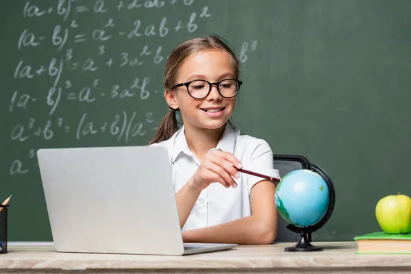 Smiling schoolkid pointing with pencil at globe near laptop and blurred chalkboard — Stock Photo