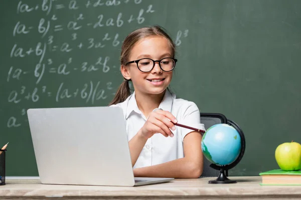 Scolaro sorridente che punta con la matita al globo vicino al computer portatile e alla lavagna sfocata — Foto stock