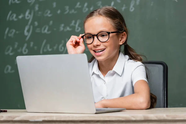 Positive schoolkid adjusting eyeglasses near laptop and chalkboard on blurred background — Stock Photo