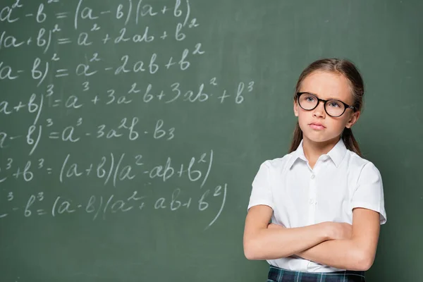 Offended schoolgirl standing with crossed arms near chalkboard with equations — Photo de stock