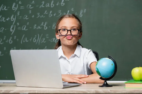 Playful schoolgirl with pencil between lips and nose sitting near laptop, globe and blurred chalkboard — Stock Photo
