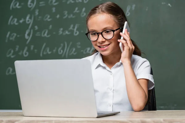 Улыбающийся школьник разговаривает на смартфоне рядом с ноутбуком и размытая доска с уравнениями — стоковое фото
