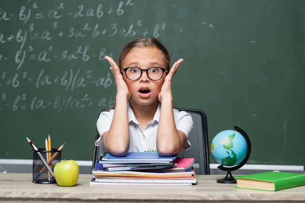 Shocked schoolkid with open mouth looking at camera near notebooks and blurred chalkboard — Photo de stock