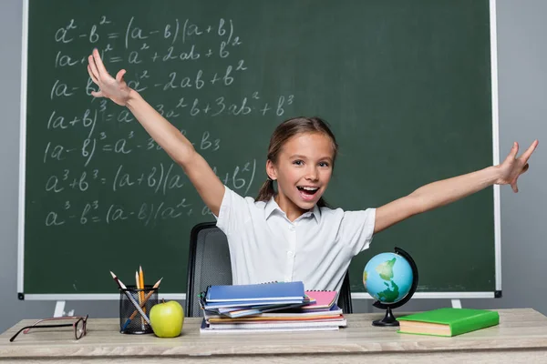 Cheerful schoolgirl with open arms near notebooks on desk and chalkboard with equations - foto de stock