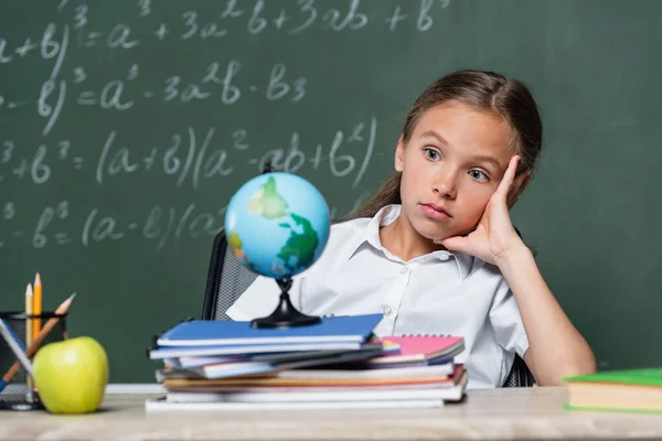 Thoughtful schoolgirl looking at globe near notebooks and chalkboard on blurred background — Photo de stock