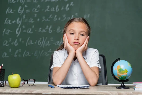 Upset schoolkid sitting at desk near globe, notebook and apple — Stock Photo
