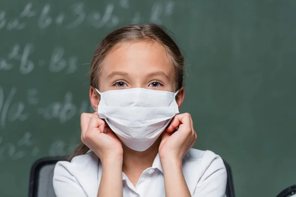 Preteen schoolgirl in protective mask looking at camera near blurred chalkboard on background - foto de stock