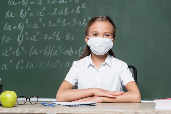 Schoolkid in medical mask sitting at desk near apple, notebook, eyeglasses and blurred chalkboard — Photo de stock
