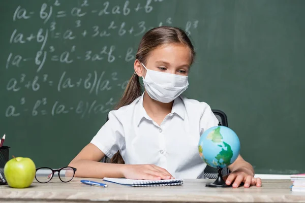 Schoolgirl in medical mask looking at globe near notebook, eyeglasses, apple and chalkboard on blurred background — Photo de stock