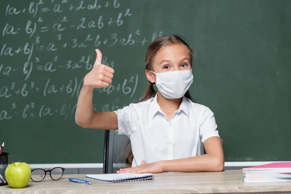 Schoolkid in medical mask showing thumb up near apple, notebook and chalkboard on blurred background - foto de stock