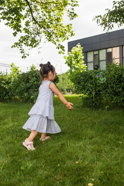 Asian girl in dress walking on grass and looking at soap bubble outside — Stock Photo