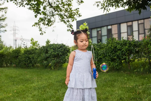 Ásia menina no vestido segurando bolha vara e olhando para sabão bolha fora — Fotografia de Stock