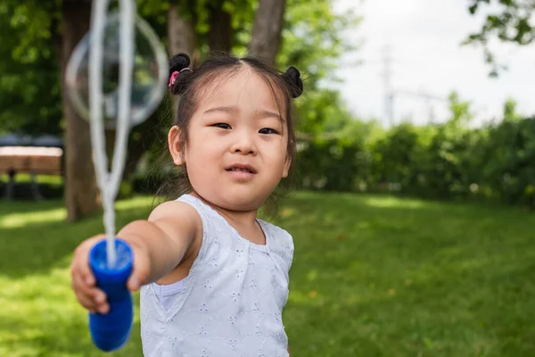 Asiatische Mädchen im Kleid hält verschwommenen Bubble Stick in der Nähe Seifenblase außerhalb — Stockfoto