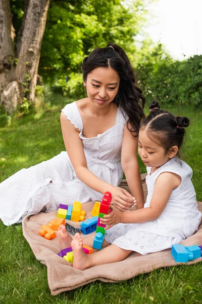 Счастливая азиатская мать смотрит на малыша дочь играет строительные блоки на одеяло для пикника в парке — стоковое фото