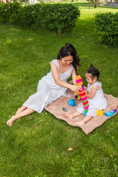 Alto ángulo vista de feliz asiático madre y niño niña jugando bloques de construcción en picnic manta en parque - foto de stock