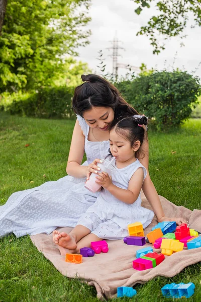 Cheerful asian mother looking at toddler girl in dress drinking milkshake near building blocks on picnic blanket — Stock Photo