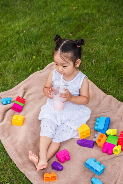 Alto ángulo vista de asiático niño niña en vestido beber sabroso batido cerca de bloques de construcción en picnic manta en parque - foto de stock
