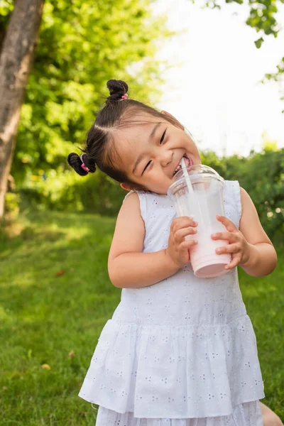 Счастливая азиатская малышка в платье пьет вкусный молочный коктейль в парке — стоковое фото