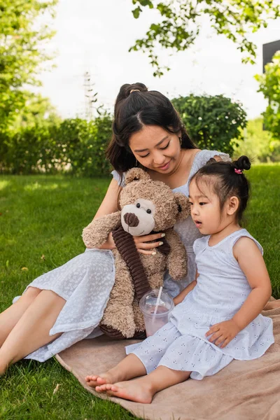 Gai asiatique mère tenant teddy barbe et regardant tout-petit fille en robe tenant milkshake sur pique-nique couverture — Photo de stock