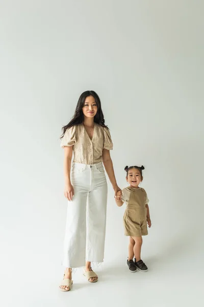 Piena lunghezza di elegante asiatico madre holding mano con felice bambino figlia su grey — Foto stock