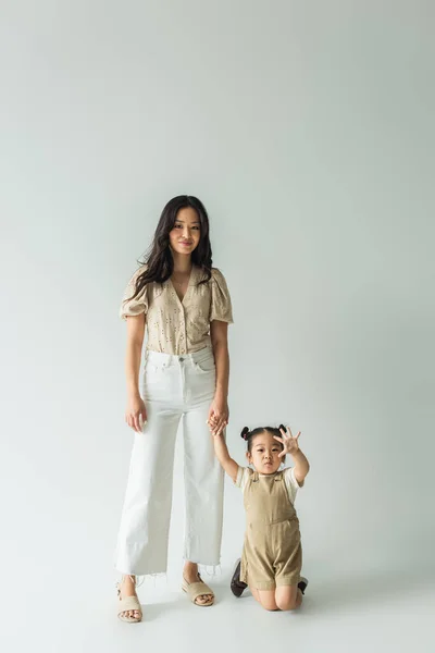 Full length of stylish smiling asian mother holding hands with toddler daughter on grey — Stock Photo