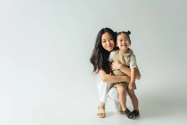 Piena lunghezza di elegante asiatico madre abbraccio bambino figlia su grigio — Foto stock