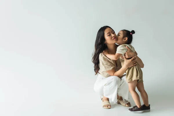 Повна довжина стильної азіатської матері обіймає дочку-малюка, цілує щоку на сірому — стокове фото