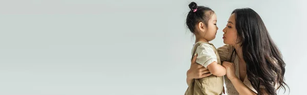 Side view of stylish asian mother hugging toddler daughter isolated on grey, banner — Stock Photo