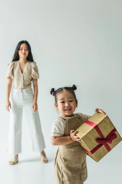 Happy asian toddler kid holding present near blurred mother on grey — Stock Photo