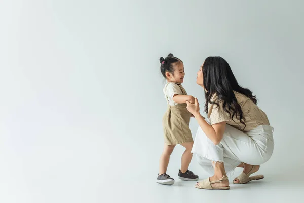 Longitud completa de feliz asiático niño pequeño cogido de la mano con elegante madre en gris - foto de stock