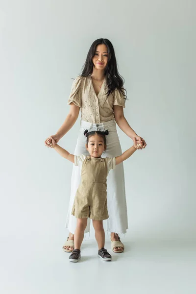 Longitud completa de feliz asiático niño pequeño y elegante madre cogido de la mano en gris - foto de stock