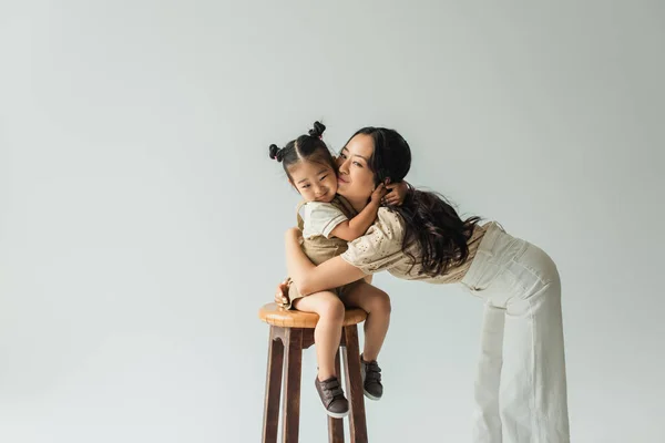 Happy asian toddler girl sitting on chair and hugging mother isolated on grey — Stock Photo