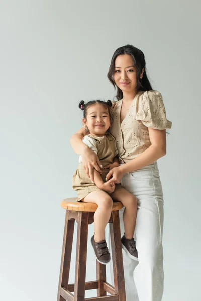Asian toddler girl sitting on chair and hugging with mother isolated on grey — Stock Photo