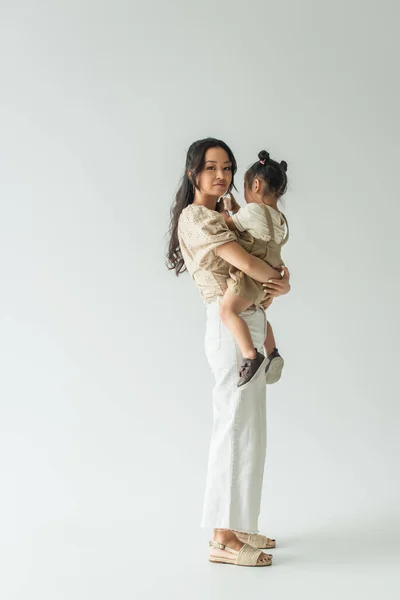 Longitud completa de feliz asiático mujer holding en brazos niño hija en gris - foto de stock