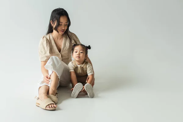 Full length of asian mother sitting with toddler daughter on grey — Stock Photo